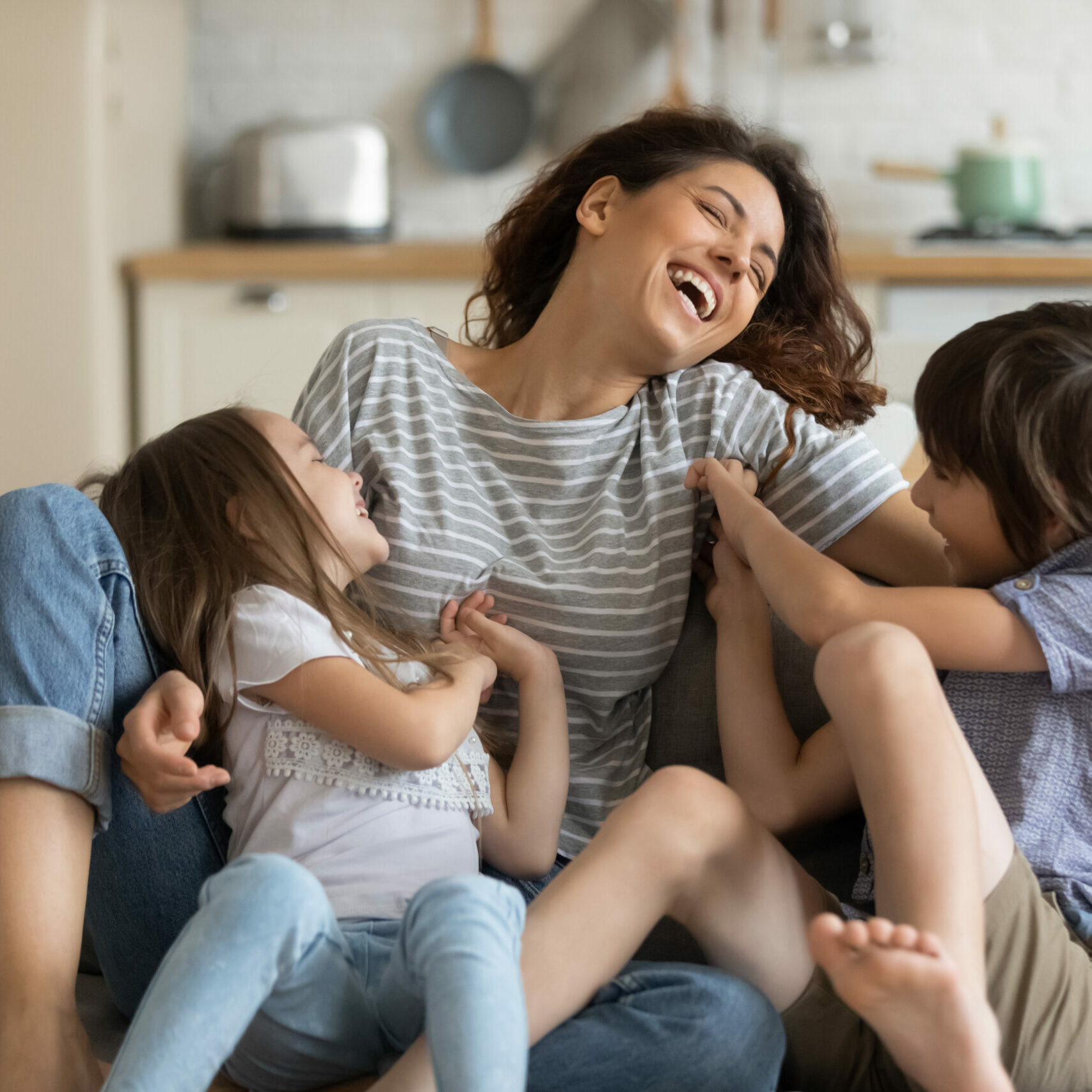 Happy cute daughter and son tickling young attractive mother sitting on couch in living room. Close up family having fun at home. Parents playing with cute brother and sister.